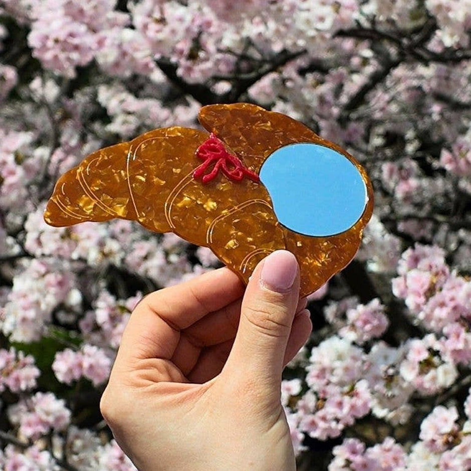 Hand-painted bow croissant compact mirror held against blooming pink flowers