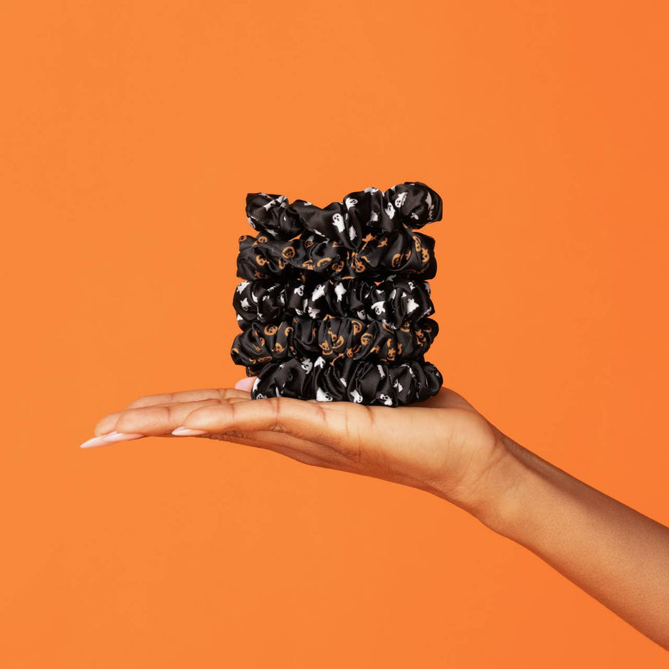 Hand holding black satin scrunchies with ghost and pumpkin Halloween print against orange background
