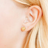 Gold shell stud earrings worn by woman with blonde hair, shown on ear in natural light