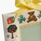Rifle Paper Co. Baby's First Christmas Picture Frame Ornament with embroidered holiday designs and yellow ribbon