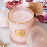 Pink candle with a gold lid and label, surrounded by blurred product images in the background.