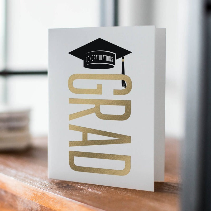 Congratulations Grad greeting card with gold text and mortarboard design on wooden surface