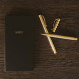 Black NOTES booklet with gold pens arranged on dark wood surface, styled desktop setting