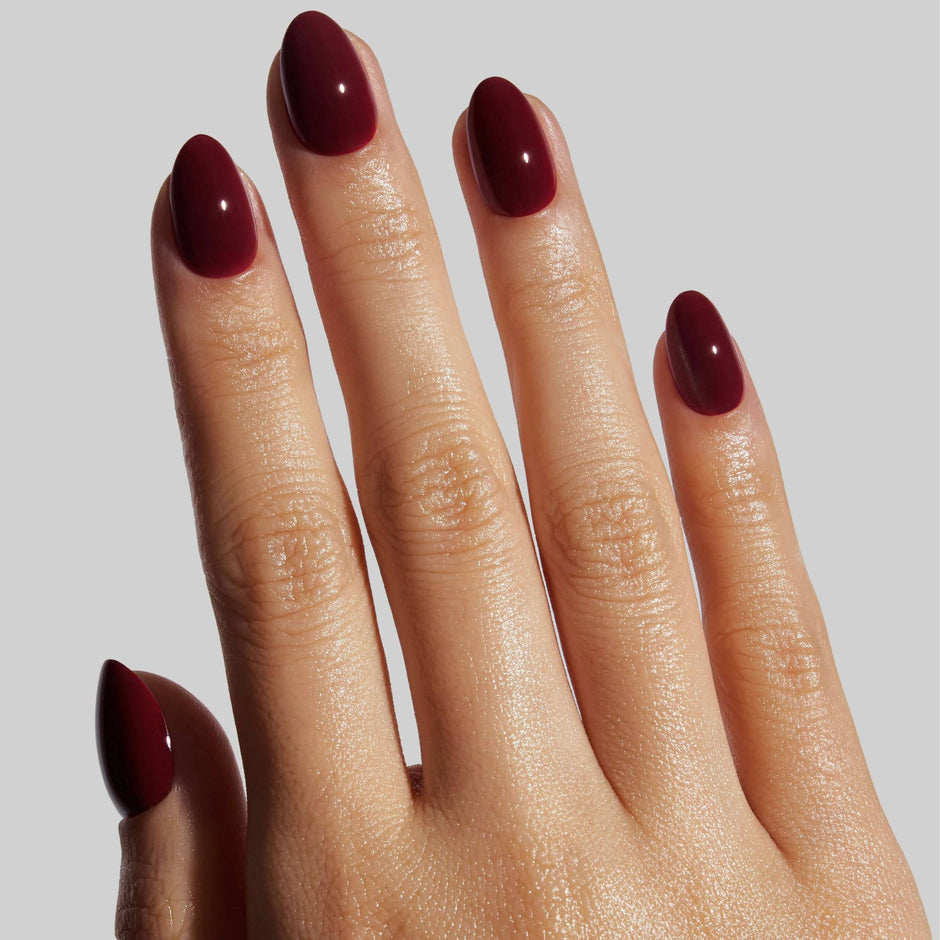 Hand displaying burgundy almond-shaped press-on nails with glossy finish against neutral background