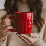 Woman holding red cable knit coffee mug with cozy sweater, wearing gold ring