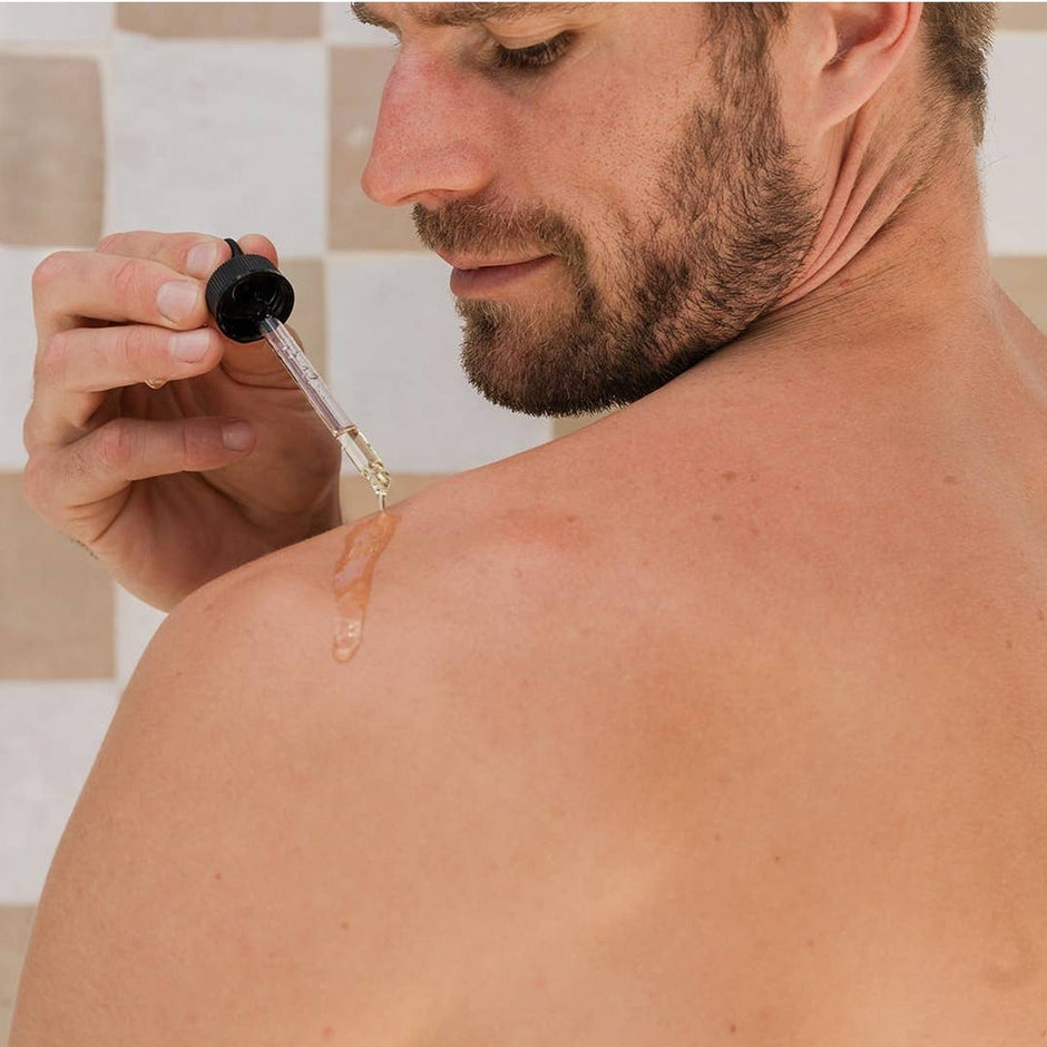 Man applying dry body oil to shoulder in bathroom, demonstrating product use