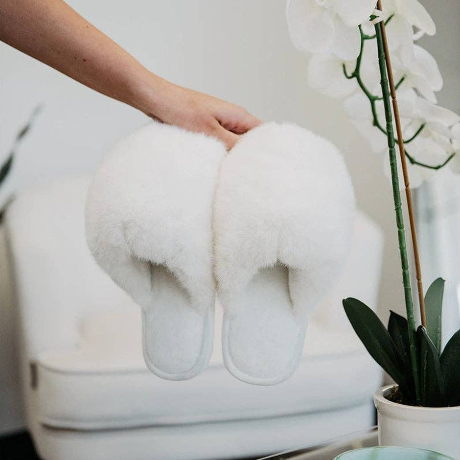 White faux fur jumbo slippers held by hand in a styled home setting with orchids and plants
