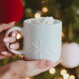 White stoneware snowflake mug with whipped cream topping, held in hands near holiday lights