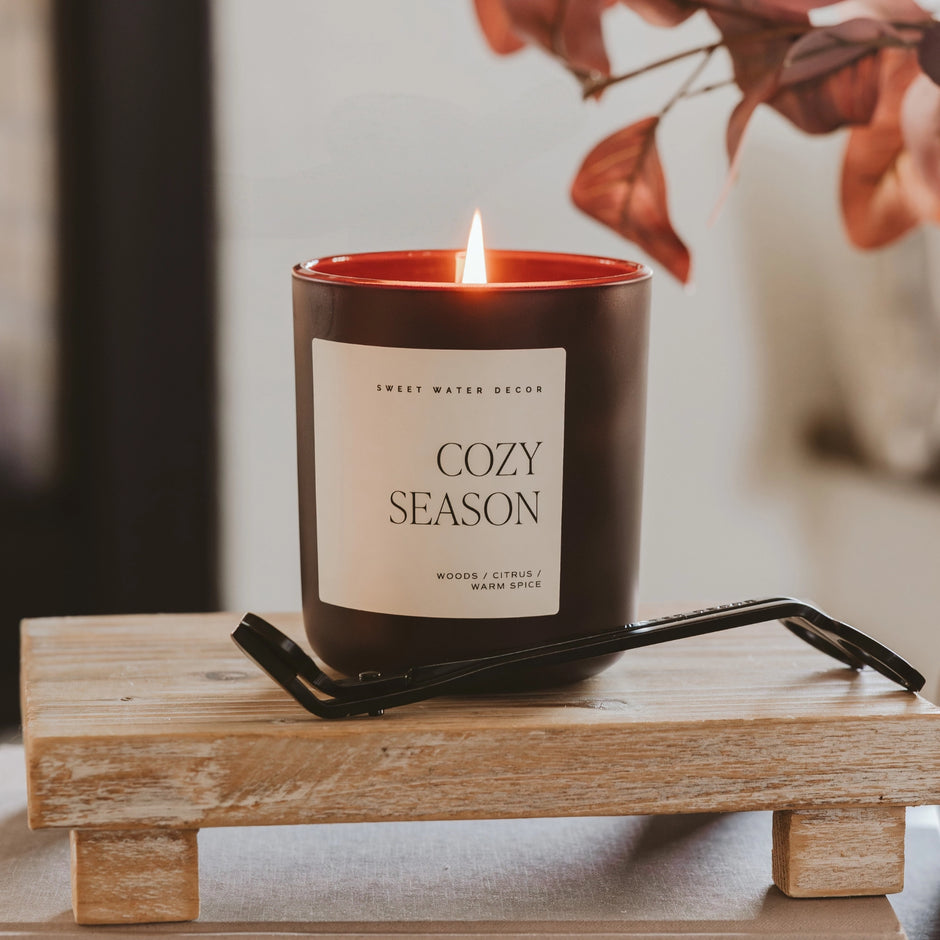 Candle labeled 'Cozy Season' on a wooden stand with a blurred background