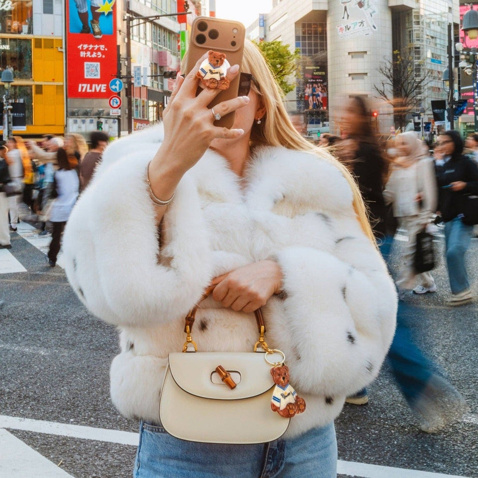 Hand-painted teddy bear bag charm attached to cream handbag, worn by woman in white fur jacket at busy city street