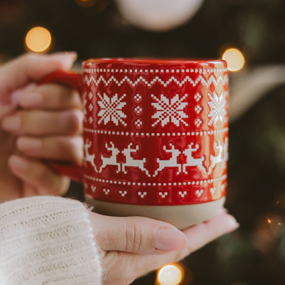 Red Fair Isle sweater stoneware mug held in hands with white sleeve, holiday lights background
