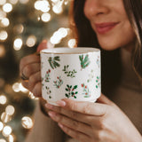 Woman holding Holiday Greenery Coffee Mug with festive botanical design and twinkling lights background