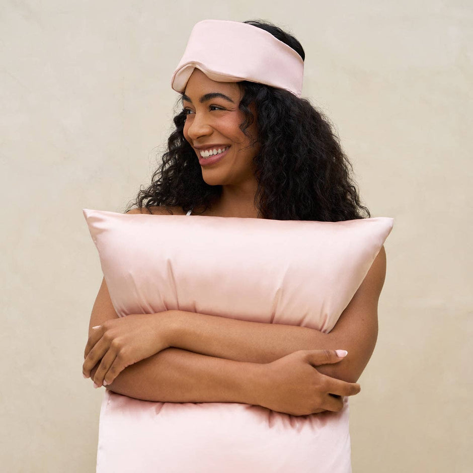 Woman wearing pink eye mask and visor, holding matching pillow, smiling against neutral background