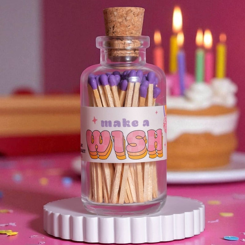 Decorative match bottle with purple tips displayed on pink surface with birthday cake and lit candles