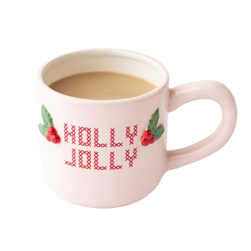 Pink ceramic mug with 'Holly Jolly' cross-stitch design and red berry decorations, filled with coffee