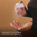 Hand holding Touchland Power Mist hand sanitizer bottle against warm tan background
