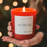 Ho Ho Ho red soy candle by Sweet Water Decor held in hands with holiday bokeh background