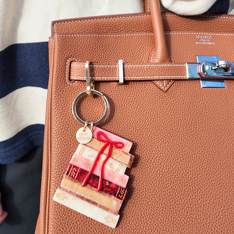 Hand-painted bow bookstack bag charm attached to a tan leather Hermès handbag, worn by a person in navy and white striped clothing