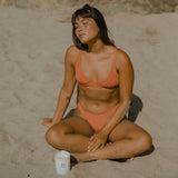 Woman in an orange bikini sitting on a sandy beach with a blurred natural background