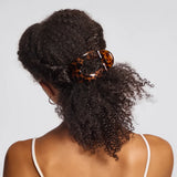 A woman shown from the back wearing a large tortoiseshell hair claw clip holding her curly dark hair in a low ponytail, against a light gray studio background.