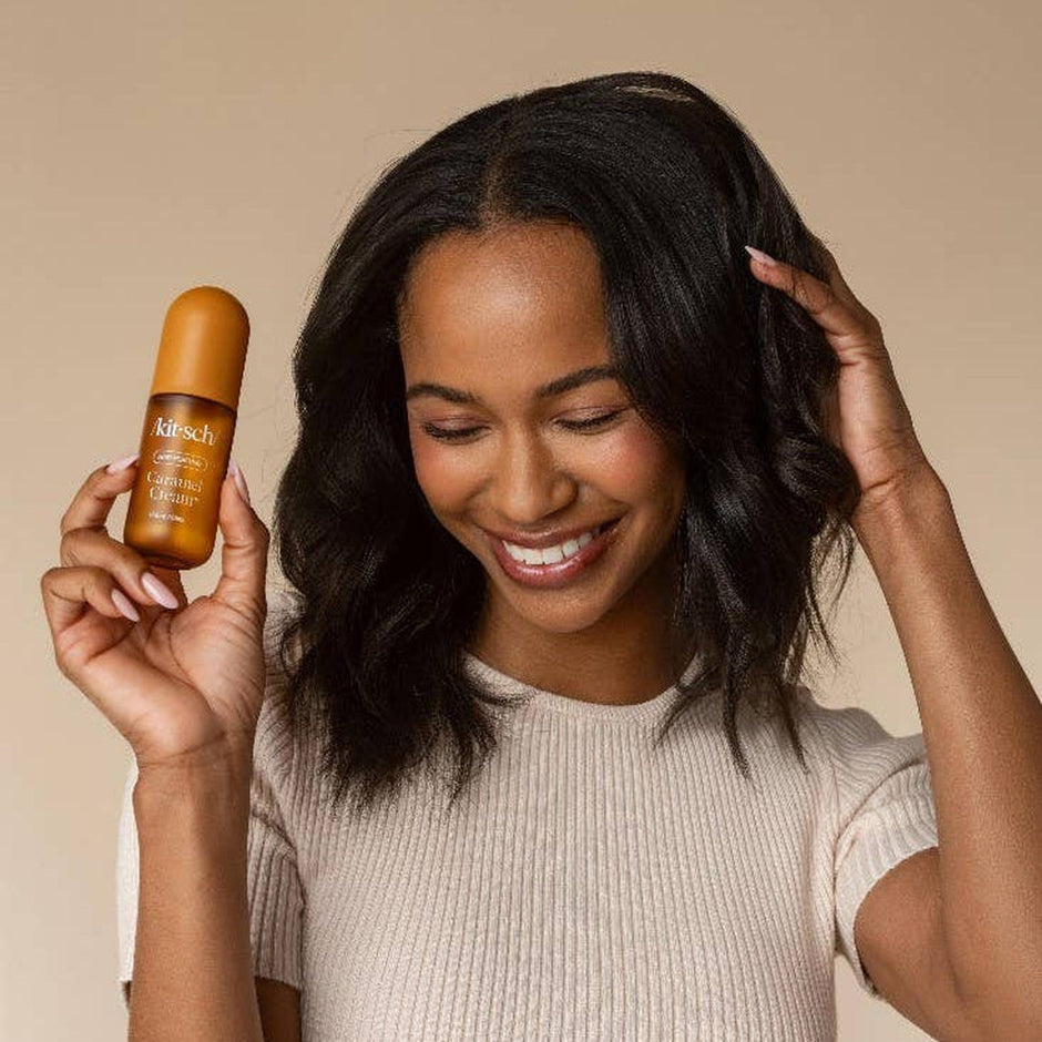 Woman holding Kitsch Caramel Cream Hair & Body Perfume Mist bottle, smiling against neutral background