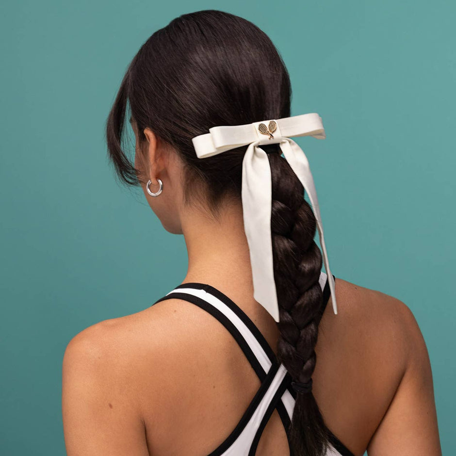 Cream satin bow hair clip worn in dark braided ponytail on model against teal background