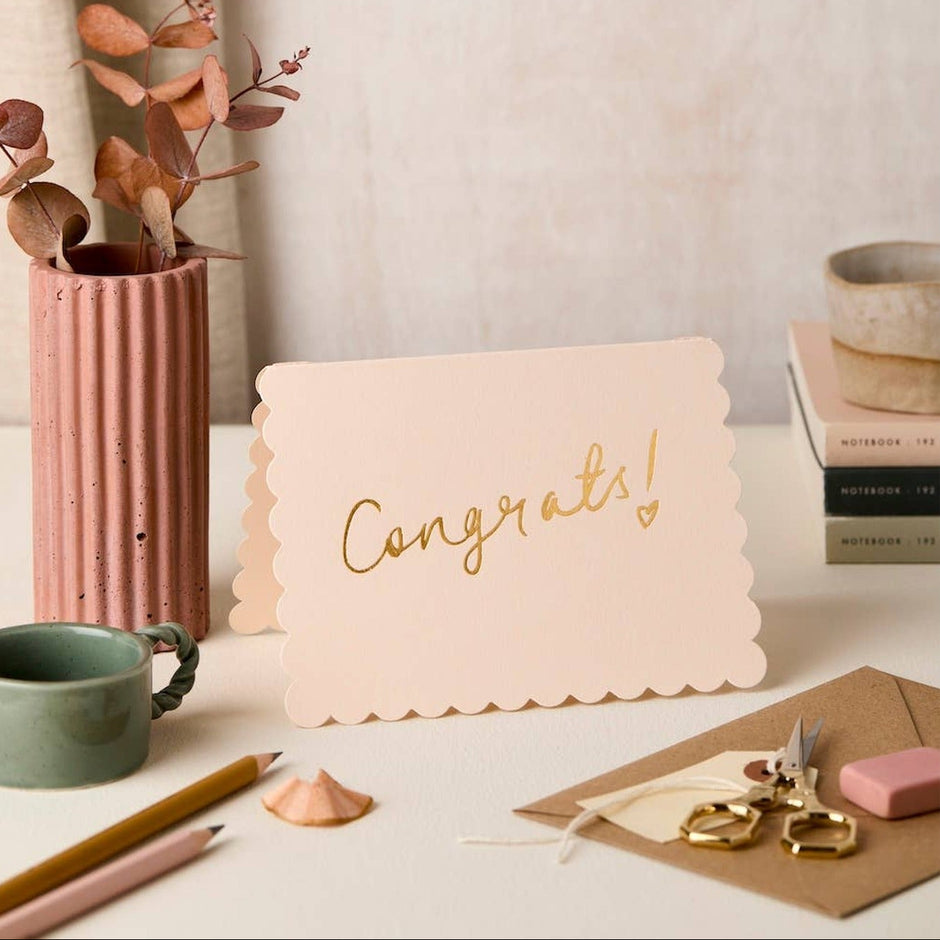 Blush scallop congratulations card displayed in styled desk setting with dried flowers and stationery