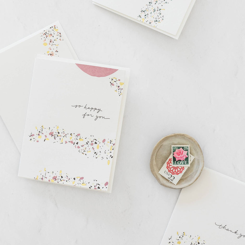 So Happy For You greeting card with terrazzo design, envelope, and love stamps on white surface