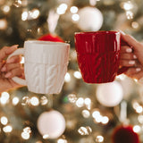 Red and white cable knit sweater mugs held by hands against festive Christmas tree background with lights