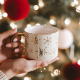 Gold snowflake coffee mug held in hands with holiday decorations and warm lights