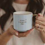 White coffee mug with 'In My Engaged Era' text held by woman wearing engagement ring