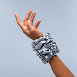 Silver satin scrunchie worn on wrist with hand raised against blue background