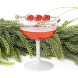 Dirty Shirley cocktail ornament with red beads, glitter details, and gold chain against evergreen branches