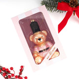 Pink teddy bear glass ornament displayed with holiday decorations including evergreen branches and red ribbon.