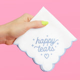 Hand holding white scalloped-edge handkerchief with blue 'happy tears' embroidery and heart design on pink background