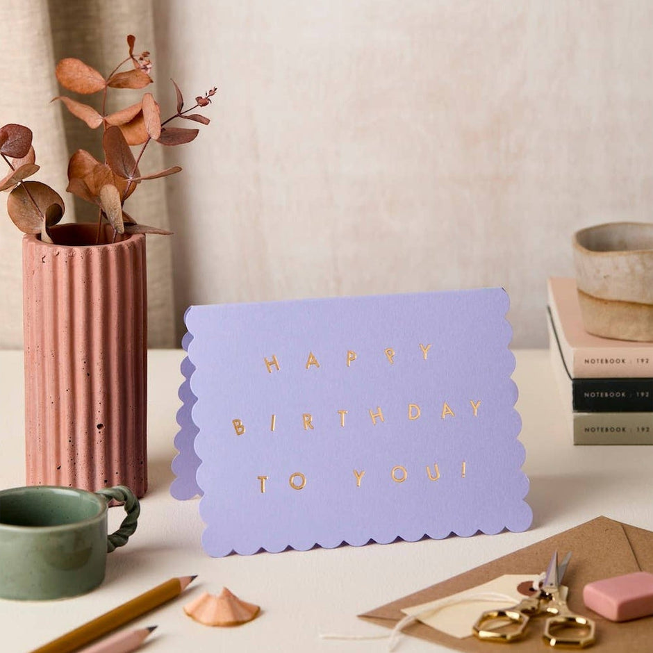 Lavender scallop birthday card displayed on desk with dried flowers, notebooks, and stationery
