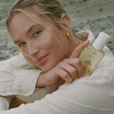 Woman holding Baja Zen In The Buff Body Glow Oil bottle in cream-colored clothing outdoors