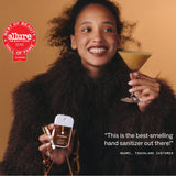 Woman holding Touchland hand sanitizer and cocktail, smiling against warm background