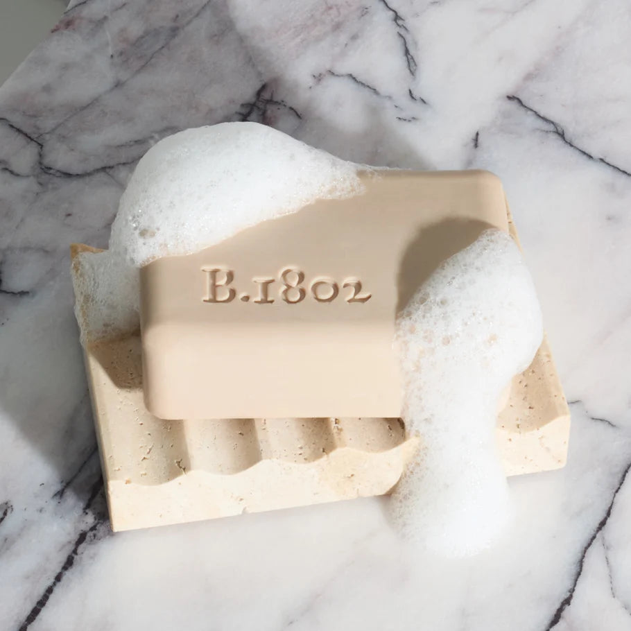 Beekman 1802 soap bar covered in lather on a travertine soap dish with a marble background