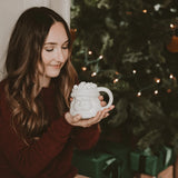 Woman holding a white ceramic mug shaped like a Santa Clause head in front of a Christmas tree.