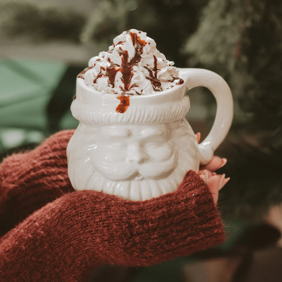 A white ceramic mug shaped like Santa's face, filled with whipped cream and drizzled chocolate, held in hands wearing a rust-brown knit sweater against a blurred holiday background.