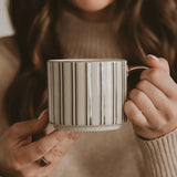 A person holding a ceramic mug with vertical green stripes on a warm beige background.