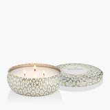 A three-wick scented candle in a round metal tin with a white and gold geometric pattern, shown next to its lid on a clean white background. The brand name VOLUSPA is visible on the lid.