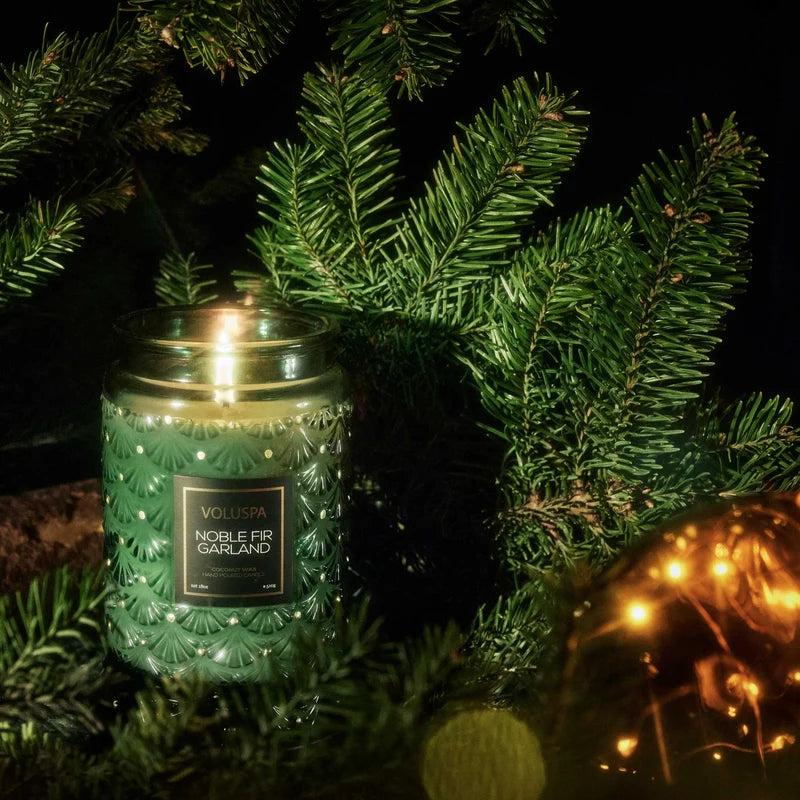 Decorative candle with a green glass jar surrounded by Christmas tree branches and ornaments.