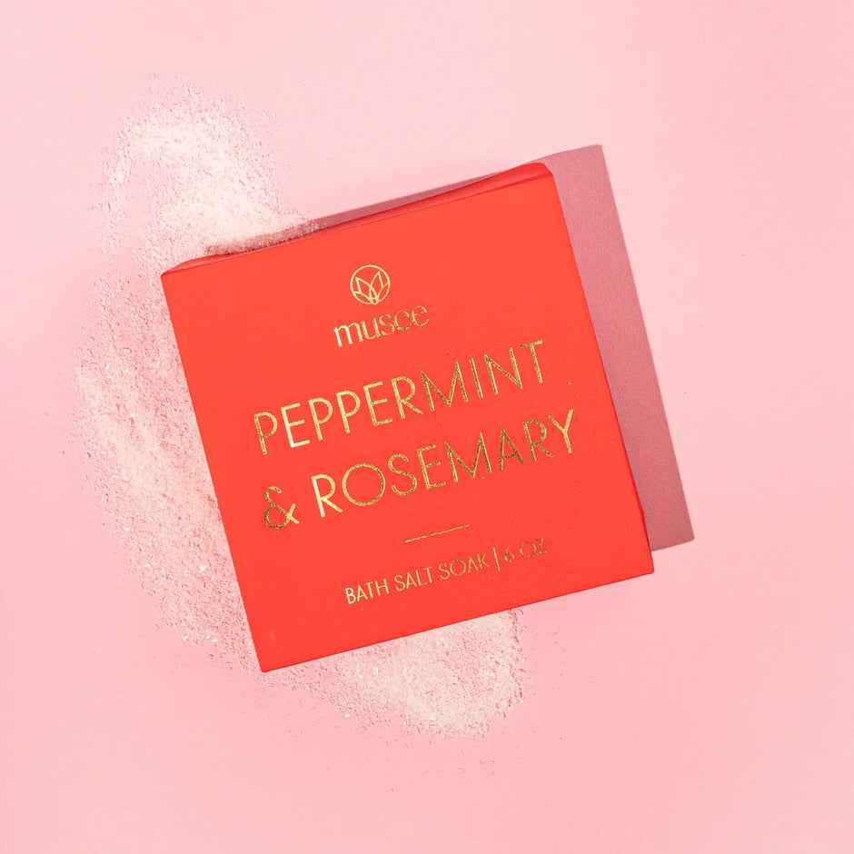 A red rectangular box of Melted Bath Salt with gold foil lettering reading 'PEPPERMINT & ROSEMARY' on a solid pink background, with white salt crystals scattered around it.