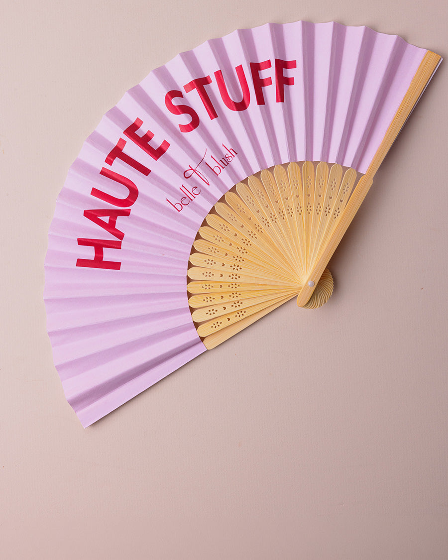 A light pink folding hand fan with a natural wood handle and ribs, featuring the words 'HAUTE STUFF' in bold red letters and 'belle & blush' in cursive. The fan is displayed against a solid beige background.