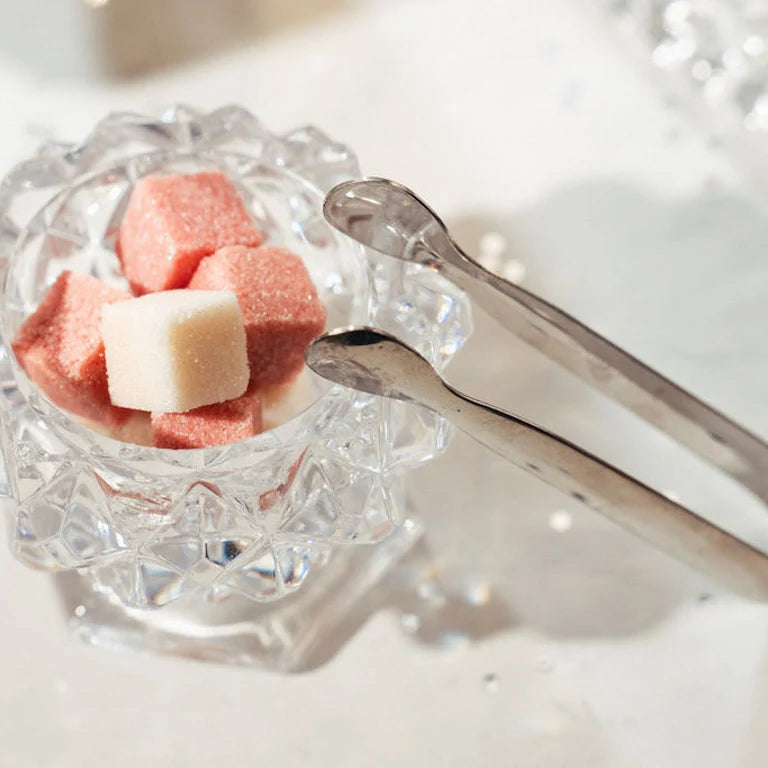 A small clear glass bowl with a faceted scalloped rim holding pink and white sugar cubes, with silver tongs resting beside it on a bright white surface.