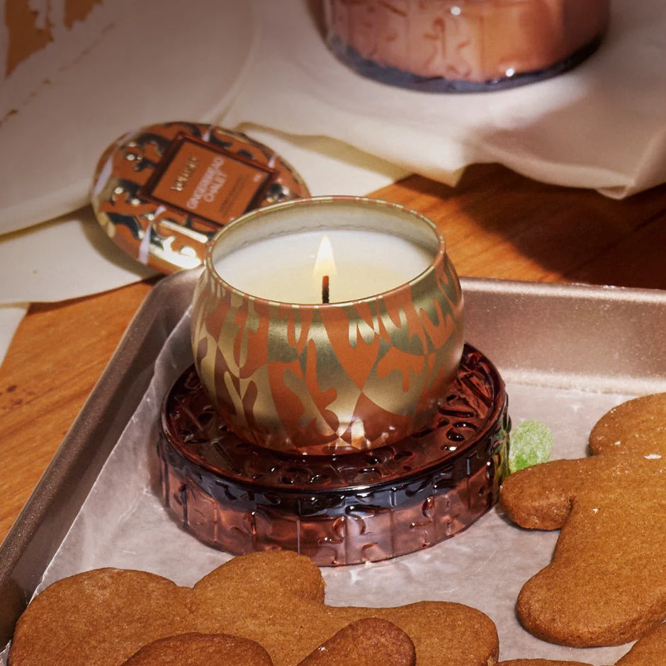 A lit scented candle in a patterned copper-toned holder sits on a tray with gingerbread cookies, with additional candles and packaging visible in a warm indoor setting.