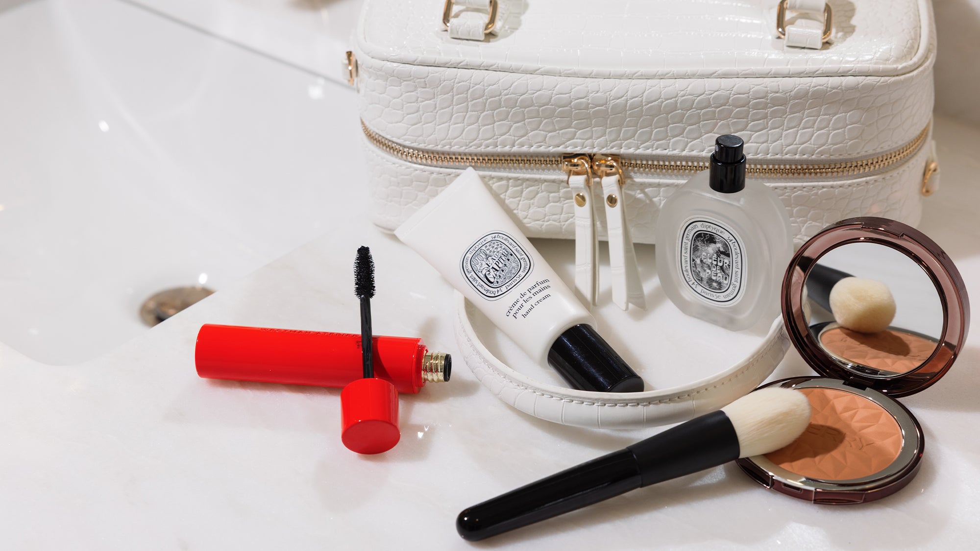 Makeup products including a brush, compact mirror, and mascara on a white surface with a white makeup bag.