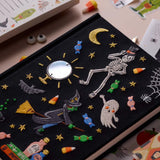 A close-up of a black fabric-covered  journal decorated with embroidered Halloween motifs, including a skeleton, moon, stars, candy, a witch riding a broomstick, a ghost, bats, and cobblestones. A small round mirror is stitched in the center.